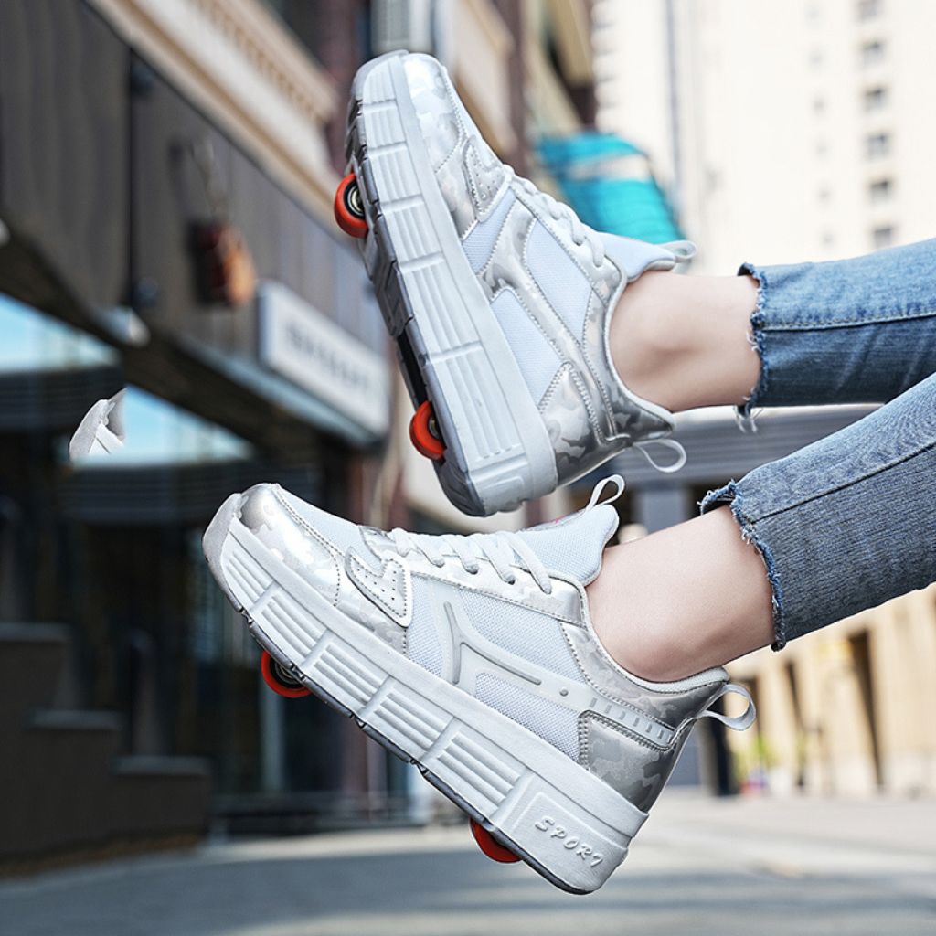 A pair of chunky platform sneakers with wheels, lace-up front, and small orange wheels attached to the sides, shown being worn with cuffed blue jeans in an outdoor urban setting
